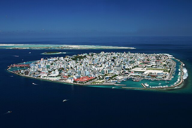 Aerial view of Malé, the capital of the Maldives, showing densely packed buildings along a low-lying coastline surrounded by turquoise ocean waters vulnerable to rising sea levels.